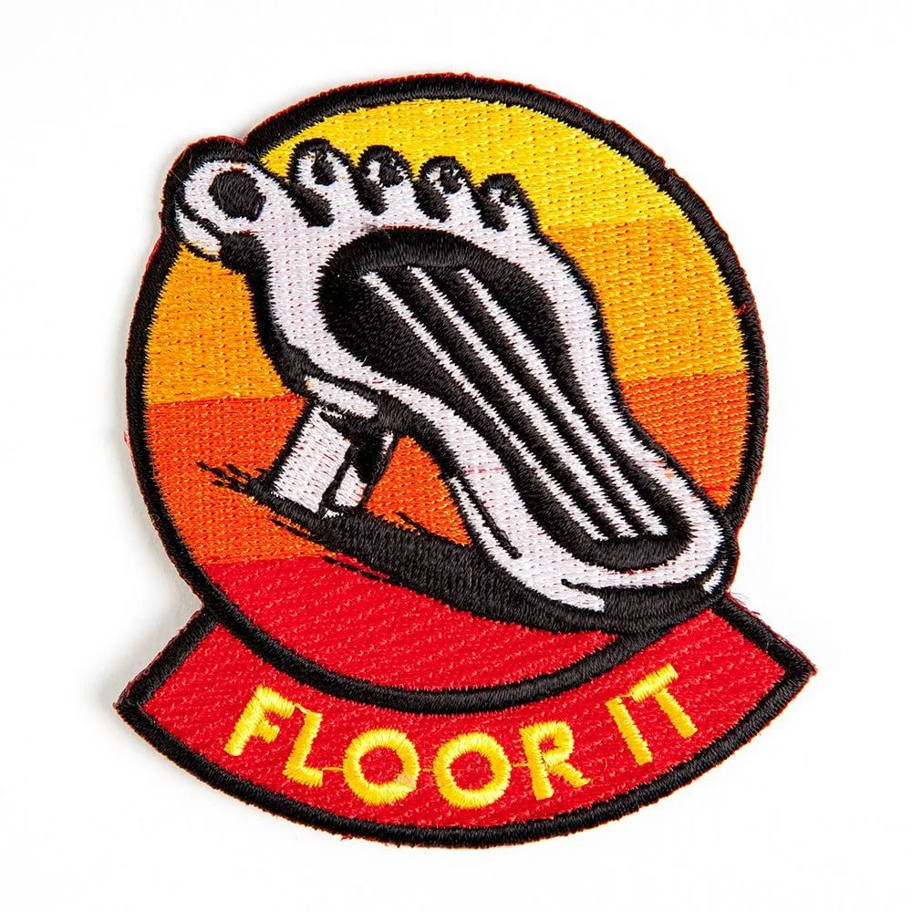 A round embroidered patch, a foot pressing a pedal against an orange sunset.