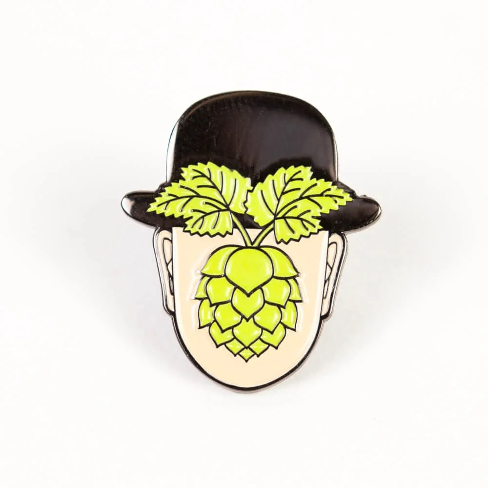 A hard enamel pin, a face under a black bowler hat with a green hop cone on it.